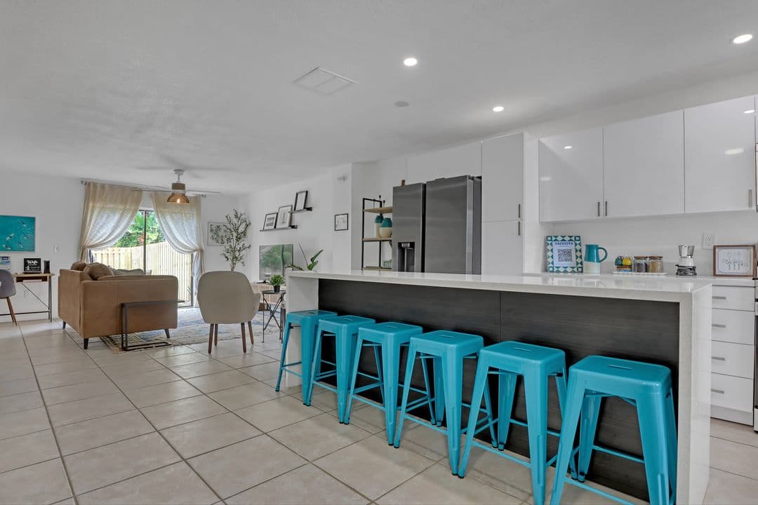 Open-concept living and kitchen area with modern white cabinets, turquoise bar stools, and natural light