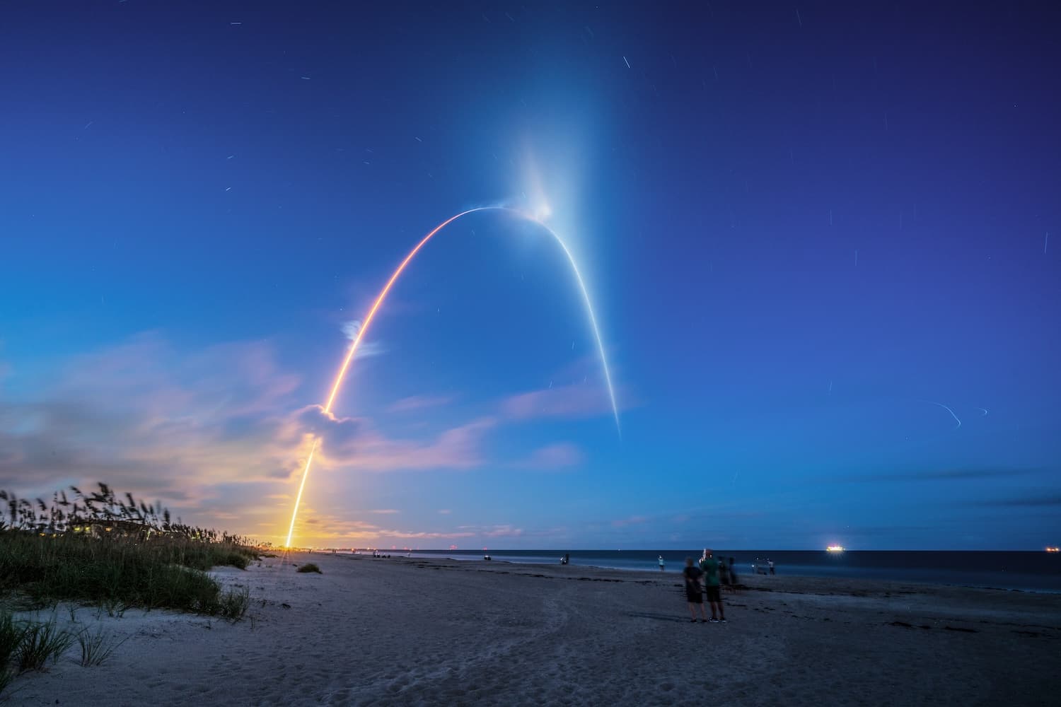 Long-exposure of a rocket launch arc over Cocoa Beach at twilight, spectators watching from the sand